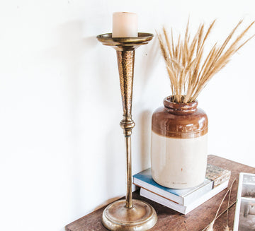 Decorative setup with a tall vintage brass candlestick, white candle, ceramic vase with dried grasses, and books on a wooden surface.