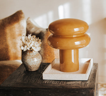 A glossy caramel orange ceramic table lamp with a rounded mushroom-shaped head and cylindrical base, displayed on a white square tray on a dark wooden side table in a warm, styled interior designer setting.