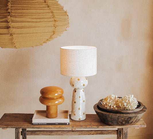 A glossy caramel orange ceramic table lamp with a rounded mushroom-shaped head and cylindrical base, displayed on a white square tray on a dark wooden side table in a warm, styled interior designer setting.