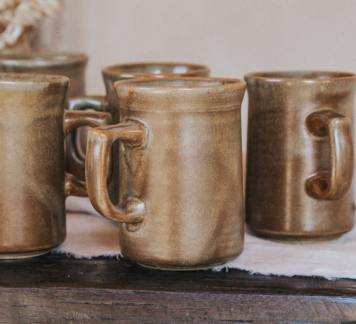 A set of rustic handmade nz temuka ceramic mugs with thick handles and a glossy earthy brown glaze, arranged on a dark wooden table over a light fabric runner.